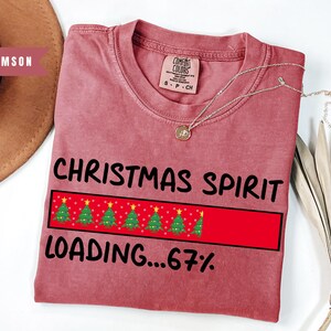 May include: Crimson-colored t-shirt with the text "CHRISTMAS SPIRIT LOADING...67%" in black. The design includes a red rectangle with Christmas trees. A silver necklace with a pendant is on the shirt.