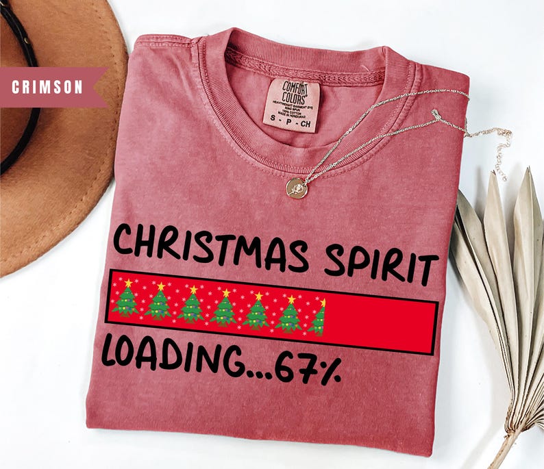 May include: Crimson-colored t-shirt with the text "CHRISTMAS SPIRIT LOADING...67%" in black. The design includes a red rectangle with Christmas trees. A silver necklace with a pendant is on the shirt.