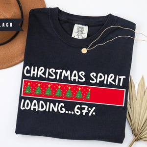 May include: Black crew neck sweatshirt with the text "CHRISTMAS SPIRIT LOADING...67%" in white. The design includes a red bar with Christmas trees. A gold necklace is draped across the sweatshirt.