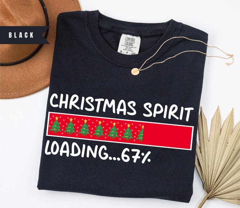 May include: Black crew neck sweatshirt with the text "CHRISTMAS SPIRIT LOADING...67%" in white. The design includes a red bar with Christmas trees. A gold necklace is draped across the sweatshirt.