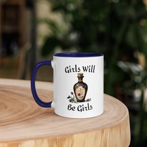 Könnte beinhalten: Eine weiße Keramiktasse mit blauem Rand. Die Tasse zeigt eine Schwarzweißillustration einer Flasche mit Blumen und dem Text "Girls Will Be Girls".