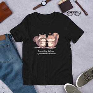 Puede incluir: Camiseta negra con un gráfico de un mapache y una ardilla tomándose de la mano. El texto "Friendship Built on Questionable Choices" está impreso debajo del gráfico.