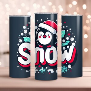 Puede incluir: Un juego de tres vasos de acero inoxidable con fondo azul oscuro y un pingüino de dibujos animados con un gorro de Papá Noel. Los vasos presentan la palabra "SNOW" en una fuente colorida y estilizada con copos de nieve y acebo.
