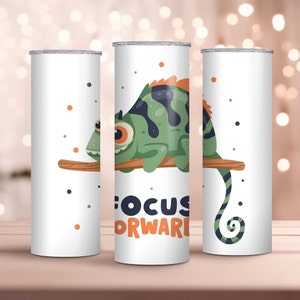 Pode incluir: Caneca branca com um camaleão de desenho animado em tons de verde, preto e laranja empoleirado em um galho marrom. A caneca tem as palavras "FOCUS FORWARD" em azul marinho e laranja. Pontos em laranja, verde e azul marinho circundam o camaleão.
