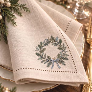 May include: A beige linen napkin featuring an embroidered wreath design with green leaves, small white berries, and a light blue ribbon bow. The napkin has a hemstitched border detail and a textured fabric weave throughout the material.