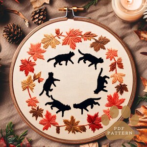 Puede incluir: Bastidor de bordado con tela crema con siluetas de gatos negros. El diseño está rodeado de hojas de otoño bordadas en tonos rojos, naranjas y marrones. El texto "PDF PATTERN" es visible.