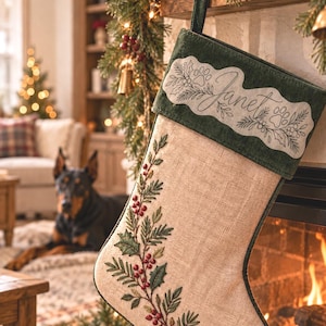 May include: A beige linen Christmas stocking featuring an embroidered holly branch with red berries and green leaves. The top cuff is dark green velvet with the name Janet embroidered in black script, surrounded by holly and paw print motifs.