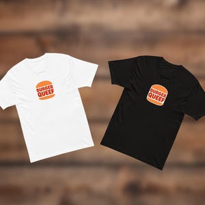 May include: A white short-sleeve t-shirt and a black short-sleeve t-shirt, each featuring a centered graphic of an orange burger icon with the text BURGER QUEEF printed in bold, dark red capital letters on the front of the chest area.