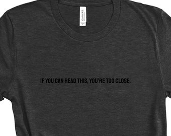 May include: A dark gray t-shirt with the text "IF YOU CAN READ THIS, YOU'RE TOO CLOSE." printed on the front.