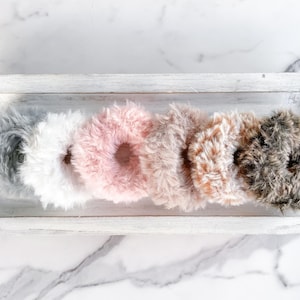 May include: Six fluffy hair scrunchies in a white rectangular tray. The scrunchies are in shades of gray, white, pink, beige, and brown.