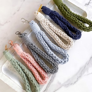 May include: A set of eight braided keychains in various colors, including green, blue, gray, pink, and beige. Each keychain has a gold metal clasp and a tassel at the end.