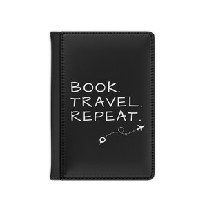 May include: Black passport holder with white text that reads "BOOK. TRAVEL. REPEAT." A dotted line with a small airplane graphic is below the text. The passport holder is rectangular and has a stitched border.
