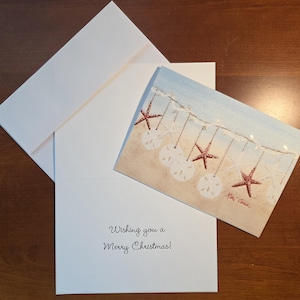May include: A Christmas card with a beach scene featuring red glitter starfish and white sand dollars hanging from a string. The card is open, revealing the message "Wishing you a Merry Christmas!" with a white envelope.