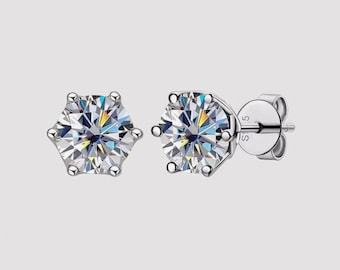 May include: A pair of stud earrings featuring a large, round, clear gemstone set in a silver-colored metal. The gemstones have multiple facets that reflect light. The earrings are displayed against a light gray background.