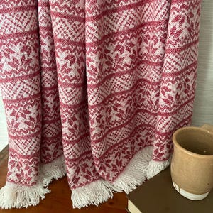May include: A red and white patterned throw blanket with a fringed edge. The blanket features a repeating geometric design. A brown mug sits on a wooden surface next to the blanket.