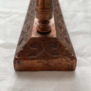 May include: Close-up of a copper-colored, rectangular object with a decorative floral pattern and a central, vertical post. The object has a weathered appearance, suggesting age or use. The base features a symmetrical design.