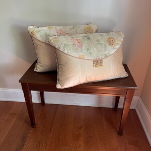 May include: Two rectangular throw pillows featuring a beige satin-like lower section and a pale green upper flap with a floral pattern in pink and yellow. The pillows are accented with twisted cord trim and a row of small yellow beaded fringe.