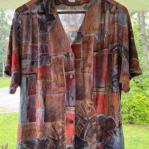 May include: A short-sleeved button-down shirt featuring a multi-colored abstract geometric and floral print in shades of brown, red, blue, and grey. The shirt has a V-neckline, a front button closure, and a label reading CASUAL CORNER.