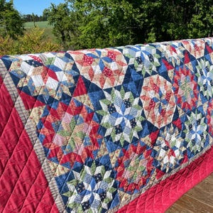 May include: A handmade quilt with a starburst pattern in red, white, blue, and green fabrics. The quilt has a red border and a striped edge. The quilt is displayed outdoors, draped over a wooden surface.