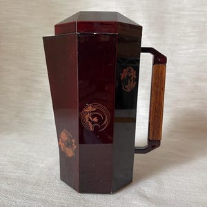 May include: A dark red, geometric-shaped pitcher with a wooden handle. The pitcher has decorative gold-colored floral designs on the front and side. The pitcher is likely made of lacquered wood.