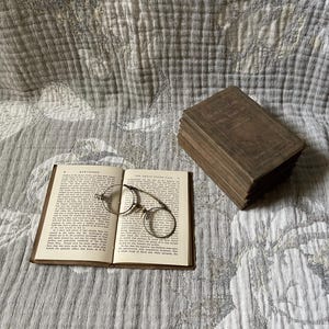 May include: An open book with text and round eyeglasses rests on a patterned surface. A stack of books is to the right. The book is open to a page with text. The eyeglasses have a thin, metal frame.