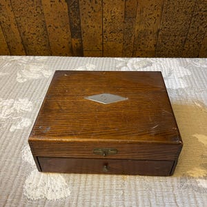 May include: A vintage wooden box with a drawer. The box is made of dark brown wood with visible grain and a small metal diamond-shaped accent on the lid. The drawer has a small brass knob. The box sits on a patterned surface.