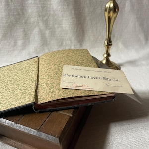 May include: An open antique book with patterned endpapers and a business card from The Bullock Electric Mfg Co. rests on a wooden box. A brass candlestick stands in the background, creating a vintage aesthetic.