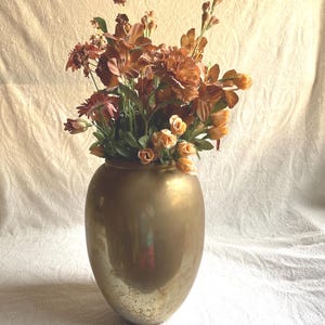 May include: A metallic gold vase with a smooth, rounded body and a slightly weathered, speckled finish at the base. The vase is filled with an arrangement of artificial flowers in various shades of orange, copper, and muted peach tones.