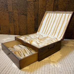 May include: A vintage wooden box with a drawer, lined with cream-colored satin fabric. The box is open, revealing the interior and drawer, suggesting storage for jewelry or keepsakes. The wood has a rich, dark finish.