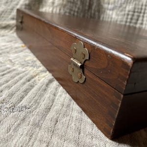 May include: A rectangular wooden box with a dark brown finish. The box has a brass-colored clasp and hinges. The box is closed and sits on a textured, light-colored fabric.