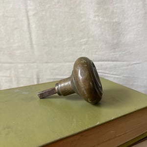 May include: An antique brass doorknob with a rounded, oval shape and a tarnished finish. The doorknob is resting on a stack of old books with green and tan covers. The doorknob's spindle is visible, suggesting it's ready for installation.