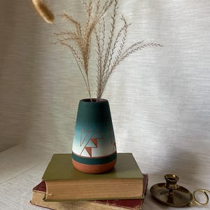 May include: A teal and white ceramic vase with a geometric design, holding dried ornamental grasses. The vase sits atop two vintage books with a brass candlestick holder nearby. The vase has a terracotta base.