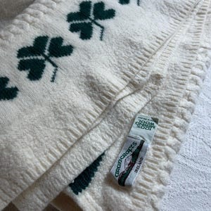 May include: An off-white knit throw blanket featuring a repeating dark green shamrock pattern and cable knit texture. A white fabric tag is attached, displaying the text Aran Crafts, 100% Merino Wool, and Made in Ireland in black lettering.