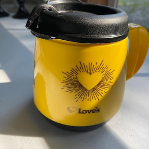 May include: A bright yellow travel mug with a black lid and handle. The front features a dark brown graphic of a heart surrounded by radiating lines, positioned above the text "Love's" with a small heart icon preceding the word in dark brown ink.