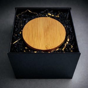 May include: A circular, light brown wooden item is presented in a black box with black and gold shredded paper. The wooden item displays a natural wood grain and is centrally placed. The square box has a matte finish.