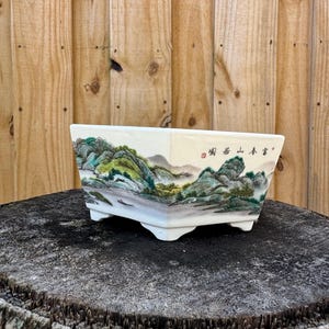 May include: Square ceramic planter with a painted landscape scene of mountains and trees. The planter has a white background with green, brown, and blue accents. The text "寫意 山水" is written in black on the planter.