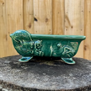May include: A green ceramic bathtub planter with a dragon design. The planter has three legs and is sitting on a wooden surface.