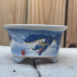 May include: A small, square ceramic planter with a blue and white dragon design. The dragon is depicted with yellow accents against a backdrop of clouds. The planter has three small feet and a decorative rim.
