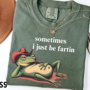 May include: A moss green t-shirt featuring a graphic of a green frog wearing a red cowboy hat and reclining. The text "sometimes i just be fartin" is printed in white above the frog. The shirt has a Comfort Colors brand label at the neck.