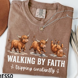 May include: An espresso-colored crew neck t-shirt featuring a graphic of three Highland cows in various poses. The text reads "WALKING BY FAITH" in large white letters, followed by "tripping constantly" in a smaller white script font.