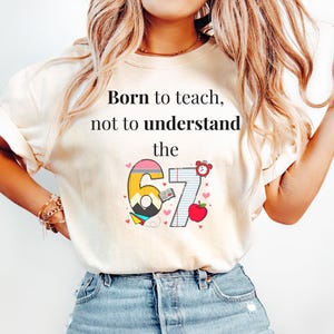 Puede incluir: Camiseta color crema con el texto "Born to teach, not to understand the" sobre un gráfico colorido de un lápiz, los números 6 y 7, un reloj y una manzana. El gráfico incluye corazones.