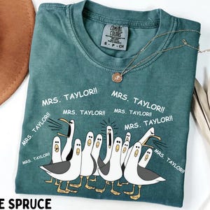 May include: Blue spruce t-shirt with a crew neck and short sleeves. The shirt features a graphic of cartoon seagulls and the text "MRS. TAYLOR!!" in white. A gold necklace with a pendant is draped on the shirt.