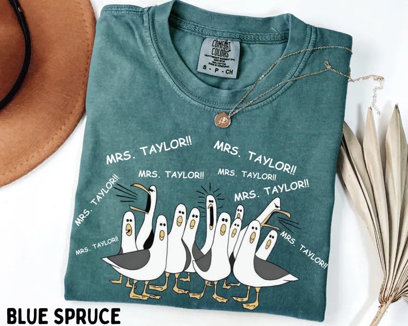 May include: Blue spruce t-shirt with a crew neck and short sleeves. The shirt features a graphic of cartoon seagulls and the text "MRS. TAYLOR!!" in white. A gold necklace with a pendant is draped on the shirt.