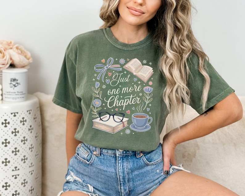 May include: Olive green t-shirt with the text "Just one more chapter" surrounded by illustrations of books, glasses, a coffee cup, and floral elements. The design features a whimsical, hand-drawn style, perfect for book lovers.