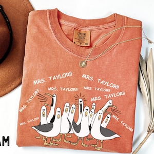 May include: Orange Comfort Colors t-shirt with a cartoon seagull design and the text "MRS. TAYLOR!!". The shirt has a crew neck and short sleeves. A gold necklace is draped across the shirt.