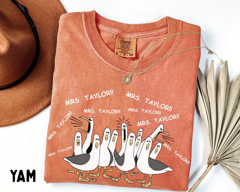May include: Orange Comfort Colors t-shirt with a cartoon seagull design and the text "MRS. TAYLOR!!". The shirt has a crew neck and short sleeves. A gold necklace is draped across the shirt.