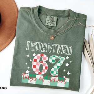 May include: A moss green t-shirt with the text "I Survived 67 Days of School" in white, red, and green. The number "67" is designed with a checkered pattern. A gold necklace with a pendant is draped on the shirt.