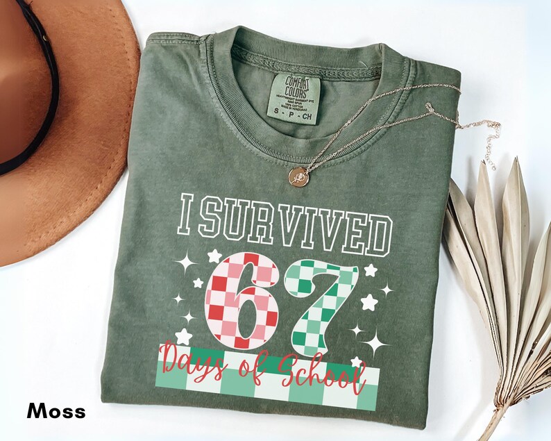 May include: A moss green t-shirt with the text "I Survived 67 Days of School" in white, red, and green. The number "67" is designed with a checkered pattern. A gold necklace with a pendant is draped on the shirt.