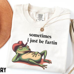 May include: An ivory t-shirt featuring a graphic of a green frog wearing a red cowboy hat and reclining. The text above the frog reads, sometimes i just be fartin. The shirt has a Comfort Colors label at the neck and a gold-toned pendant necklace.