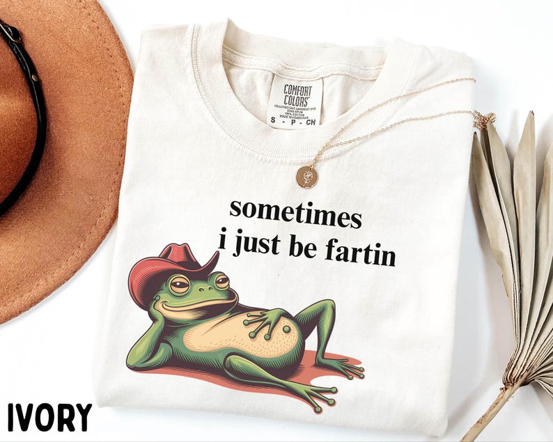May include: An ivory t-shirt featuring a graphic of a green frog wearing a red cowboy hat and reclining. The text above the frog reads, sometimes i just be fartin. The shirt has a Comfort Colors label at the neck and a gold-toned pendant necklace.